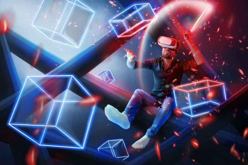 Man with red light saber sitting in blue white and red colored backdrop of geometric boxes type metaverse with headset on