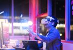 Engineering the metaverse with a Young asian male wearing VR headset, working with a 3D Augmented Reality floating interface in an office at night with a purple, pink and blue back lit background