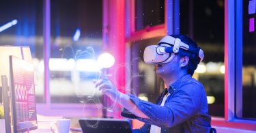 Engineering the metaverse with a Young asian male wearing VR headset, working with a 3D Augmented Reality floating interface in an office at night with a purple, pink and blue back lit background