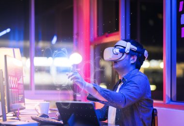 Engineering the metaverse with a Young asian male wearing VR headset, working with a 3D Augmented Reality floating interface in an office at night with a purple, pink and blue back lit background