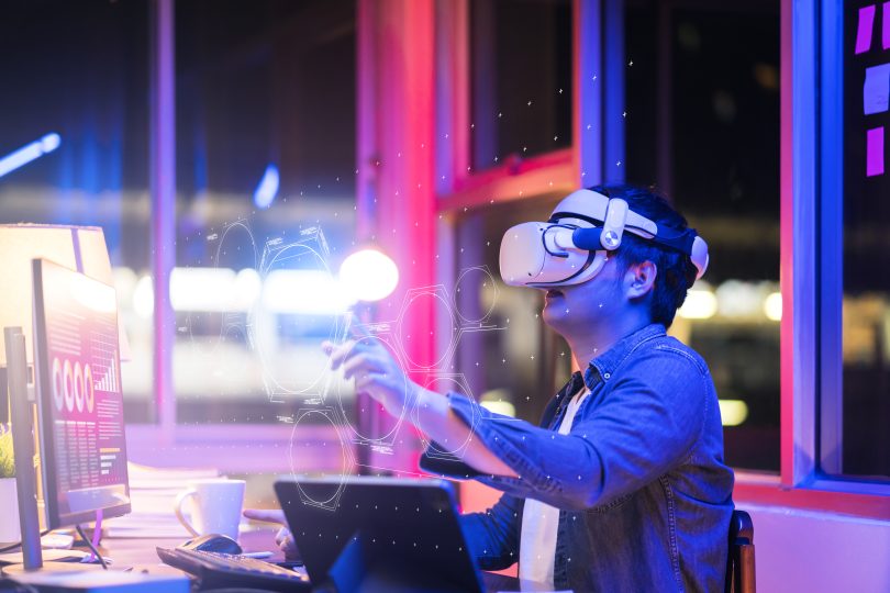 Engineering the metaverse with a Young asian male wearing VR headset, working with a 3D Augmented Reality floating interface in an office at night with a purple, pink and blue back lit background
